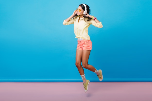 Foto gratuita foto de cuerpo entero de una niña caucásica deportiva despreocupada bailando en zapatillas de deporte. alegre modelo de mujer asiática morena en auriculares saltando, expresando emociones felices.