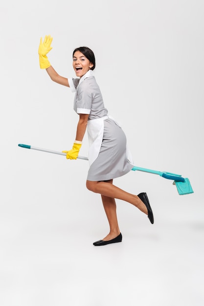 Foto de cuerpo entero de niña graciosa feliz en uniforme montando trapeador como bruja