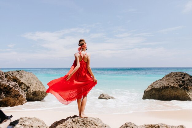 Foto de cuerpo entero de la parte posterior del modelo de mujer bonita de pie sobre una piedra grande en el océano. Retrato al aire libre de mujer delgada alegre en vestido rojo mirando al horizonte en un día ventoso.