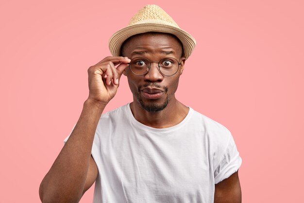 Foto de curioso hombre afroamericano se ve con expresión misteriosa, mantiene la mano en el borde de las gafas