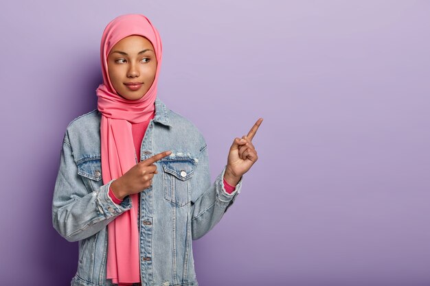 La foto de una dama árabe segura de sí misma con el tradicional hijab rosa, le muestra el camino correcto, señala el espacio en blanco con ambos dedos, invita a ir allí, promueve el espacio para copiar. Compruébalo conmigo