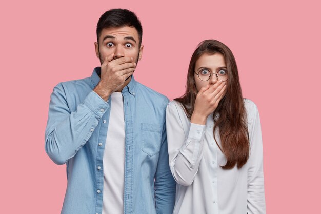 Foto de una dama y un hombre sin palabras asustados que cubren la boca con las palmas, tienen expresiones estupefactas, vestidos con una camisa elegante