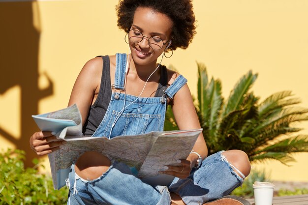 La foto de una dama negra con cabello nítido usa el mapa de destinos, busca lugares interesantes para visitar, le gusta hacer turismo en una ciudad desconocida, posa en posición de loto frente a plantas tropicales, disfruta escuchando música