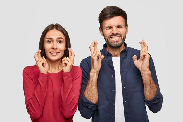 Foto de dedos cruzados femeninos y masculinos positivos para la buena suerte antes de un evento importante, parados uno al lado del otro, tener un aspecto deseable, vestido con ropa informal, aislado sobre una pared blanca.