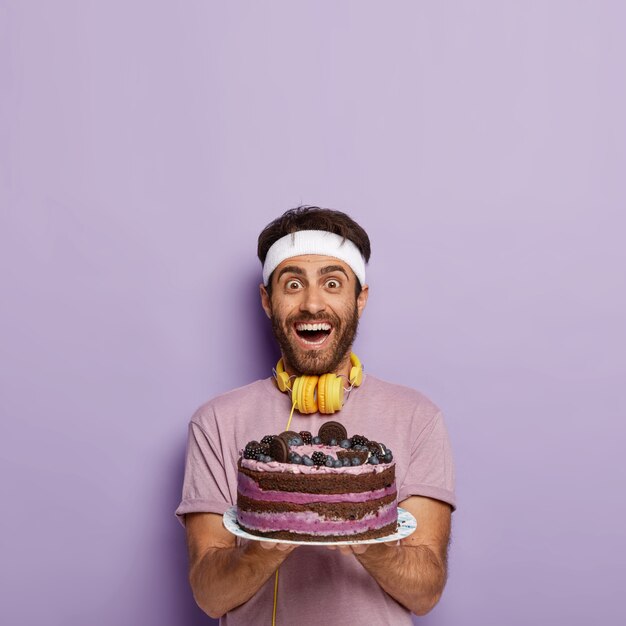 Foto de deportista feliz y lleno de alegría sostiene un gran pastel de frutas horneado con arándanos, quiere comer algo dulce después de un ejercicio agotador en el gimnasio, usa ropa informal, auriculares para escuchar audio