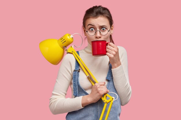 Foto de desconcertada mujer caucásica levanta las cejas, bebe bebidas de una taza roja, tiene un descanso después de estudiar, lleva lámpara de mesa