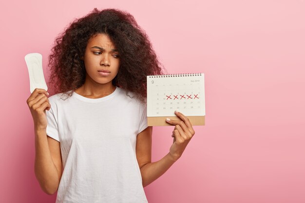 Foto de descontento encantadora mujer de pelo rizado mira el calendario con días pms marcados