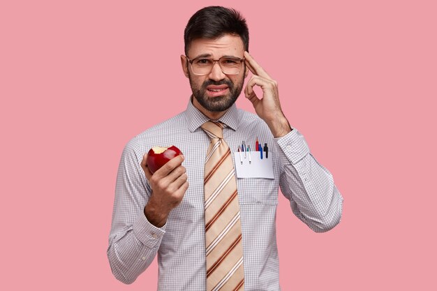 Foto de descontento joven caucásico mantiene el dedo en la sien, vestido formalmente, come manzana, recuerda algo en mente