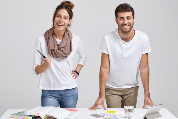 Foto de diseñadores alegres de mujeres y hombres vestidos con ropa de moda, parados cerca del escritorio blanco, estudiar literatura, hacer que el proyecto funcione en una tableta, conectado a internet inalámbrico