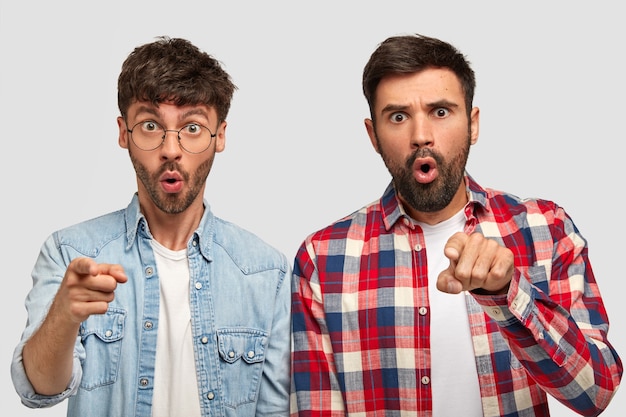 Foto de dos hombres sorprendidos con expresiones de asombro señalan con los dedos índice directamente, expresan conmoción, se paran uno al lado del otro, indican a la distancia, aislado sobre una pared blanca