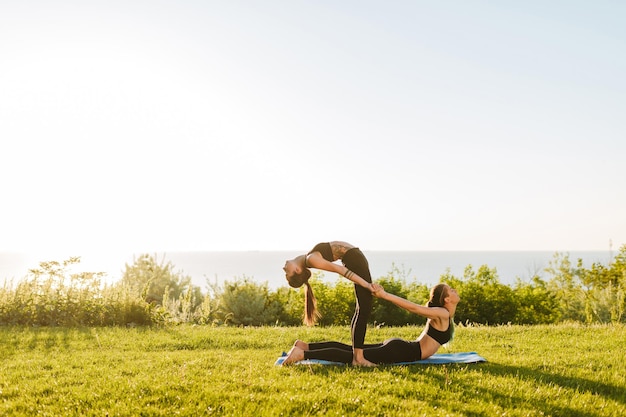 Foto de dos señoritas con tops deportivos negros y polainas entrenando poses de yoga juntas en el césped. Mujeres jóvenes practicando yoga al aire libre