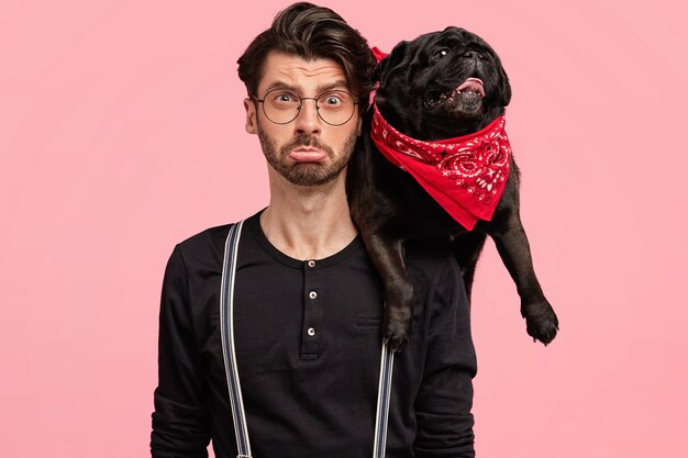 Foto del dueño masculino disgustado de perro negro de pedigrí, pasar tiempo libre en casa, aislado sobre pared rosa. Mascota divertida con pañuelo rojo en el hombro del anfitrión. Animales, familia, concepto de relación.