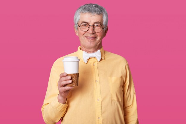 La foto de una elegante taza de café de papel para adultos mayores, lista para tomar una bebida caliente, vestida con camisa formal y corbata de moño, tiene una reunión, modelos en la pared rosada del estudio con espacio vacío para su anuncio.