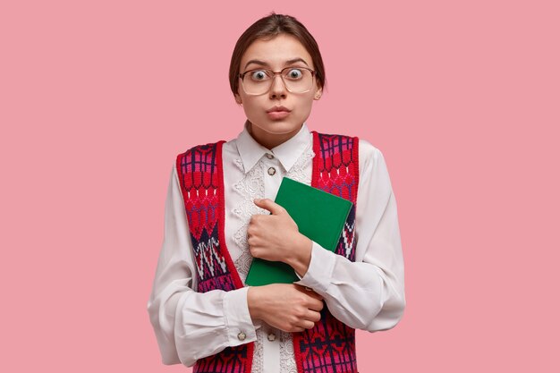 Foto de una empollona europea sorprendida que mira fijamente a través de los anteojos, viste una blusa y un chaleco formales blancos, lleva el bloc de notas verde de cerca, mucha gente sorprendida llega a la entrevista de trabajo, se preocupa antes de hablar