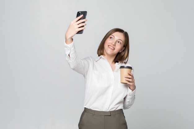 Foto de empresaria en ropa formal de pie sosteniendo café para llevar en la mano y tomando selfie en teléfono móvil sobre pared gris