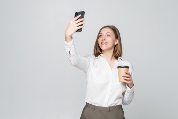 Foto de empresaria en ropa formal de pie sosteniendo café para llevar en la mano y tomando selfie en teléfono móvil