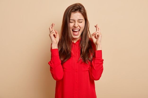 La foto de una encantadora mujer morena de pelo oscuro cruza los dedos para la buena suerte, cree sinceramente en la victoria, mantiene los ojos cerrados, vestida con una blusa roja, gestos sobre la pared beige. Lenguaje corporal