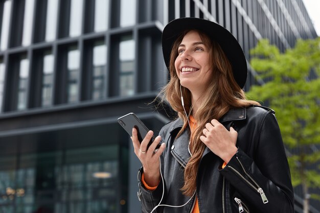 Foto de encantadora mujer soñadora sostiene un teléfono celular moderno, escucha música durante una caminata al aire libre en la metrópoli