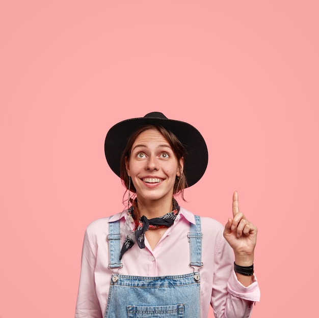 Foto de encantadora mujer tierna sonríe positivamente, mira con alegría hacia arriba, señala con el dedo índice