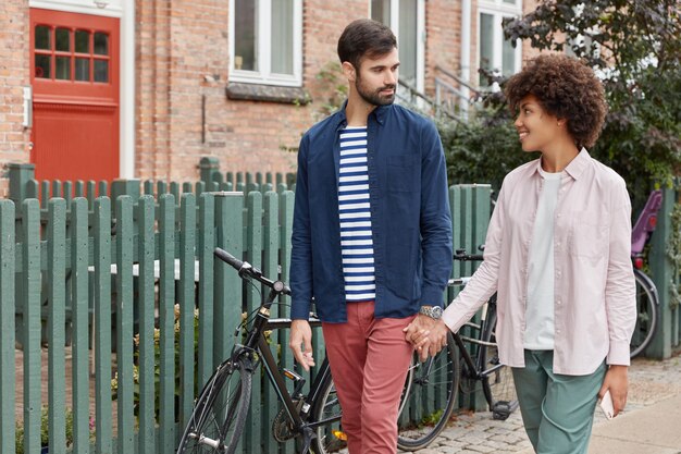 Foto de una encantadora pareja interracial caminar al aire libre, mantener las manos juntas y mirarse con amor