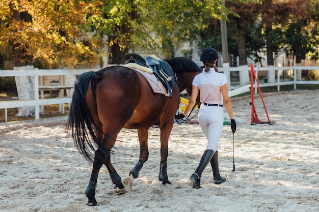 Foto de la espalda, mujer joven con uniforme especial y casco con su caballo de montar.