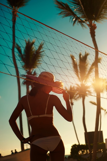 Foto en estilo retro de chica sexy modelo en bikini blanco con red de voleibol en la playa y palmeras detrás del cielo azul de verano