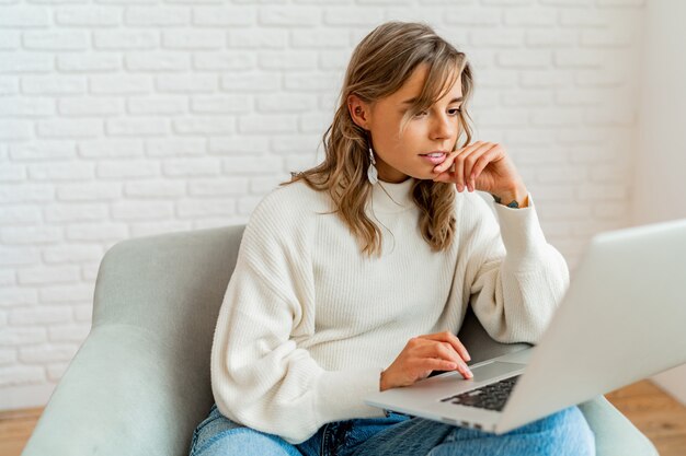 Foto de estilo de vida interior de una mujer bonita con pelos ondulados rubios sentado en el sofá en casa trabajando en la computadora portátil