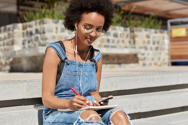 Foto de estudiante escucha audiolibro con auriculares y teléfono móvil, escribe algunos registros y detalles en el diario, posa en las escaleras al aire libre, se prepara para el seminario, usa internet y tecnología.