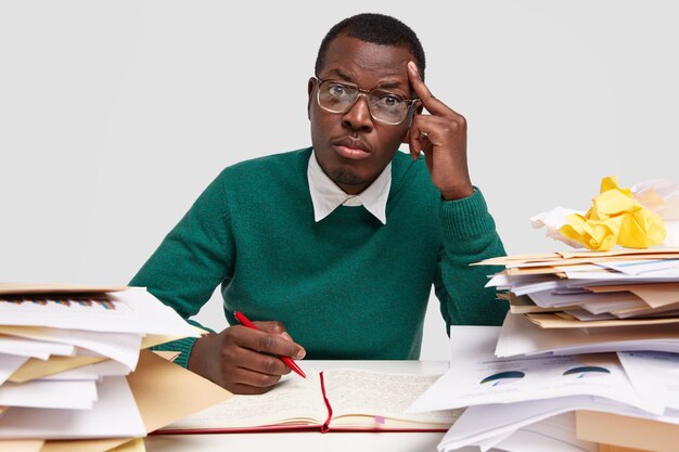 La foto de un estudiante graduado serio y elegante mantiene la mano en la sien, se sienta en el escritorio, escribe notas en el bloc de notas, usa anteojos transparentes para corregir la visión