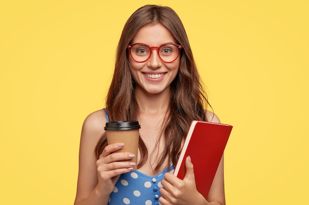 Foto de una estudiante universitaria alegre que lleva un libro de ejercicios y toma un café, sonríe ampliamente, está de buen humor después de las conferencias, se regocija en las próximas vacaciones, modelos contra la pared amarilla