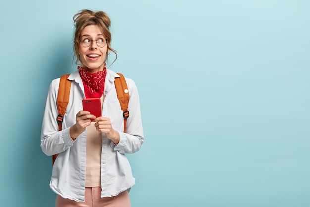 Foto de estudiante universitario alegre pensativo sostiene un dispositivo de teléfono inteligente en las manos, espera la conexión wifi, usa gafas ópticas redondas, camisa, bandana, lleva mochila. Copie el espacio en la pared azul