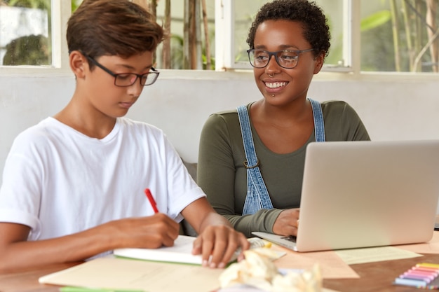 Foto de estudiantes adolescentes multiétnicos involucrados en el proceso de trabajo, buscar información en una computadora portátil portátil, escribir ideas para el trabajo del proyecto en el bloc de notas, sentarse en el escritorio, verificar el mensaje de ingresos