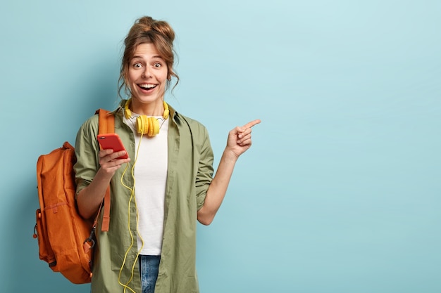 Foto de estudio de una adolescente feliz sostiene el teléfono móvil, revisa el correo electrónico, recibe una nueva notificación, usa auriculares modernos, señala el espacio de la copia para la marca o etiqueta, usa una camisa caqui suelta, lleva una bolsa