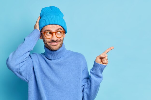 Foto de estudio de alegre adolescente masculino en ropa de moda se ve con interés y señala en el espacio de copia