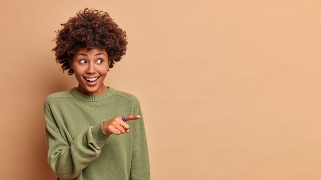 Foto de estudio de alegre mujer complacida con el pelo rizado apunta lejos en el espacio de la copia se ríe felizmente anuncia promo vestido casualmente aislado sobre pared beige