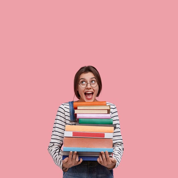 Foto de estudio de alegre mujer europea lleva gafas redondas, tiene muchos libros