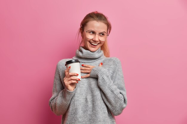 Foto de estudio de alegre niña europea ríe positivamente mantiene la mano en el pecho y sostiene el café para llevar