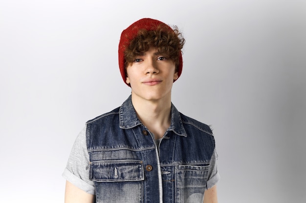 Foto de estudio de un apuesto joven de veinte años posando sobre fondo gris en blanco con espacio de copia para información vistiendo chaleco de mezclilla azul y sombrero rojo, con expresión facial segura