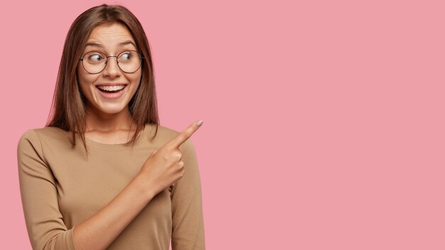 Foto de estudio de apuesto mujer morena satisfecha con amplia sonrisa dentuda, lleva gafas redondas, puntos con el dedo índice a un lado