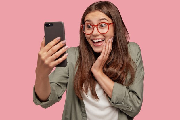 Foto de estudio de asombrada joven mujer caucásica con expresión positiva, hace selfie con teléfono celular