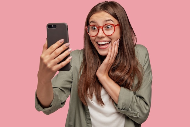 Foto de estudio de asombrada joven mujer caucásica con expresión positiva, hace selfie con teléfono celular
