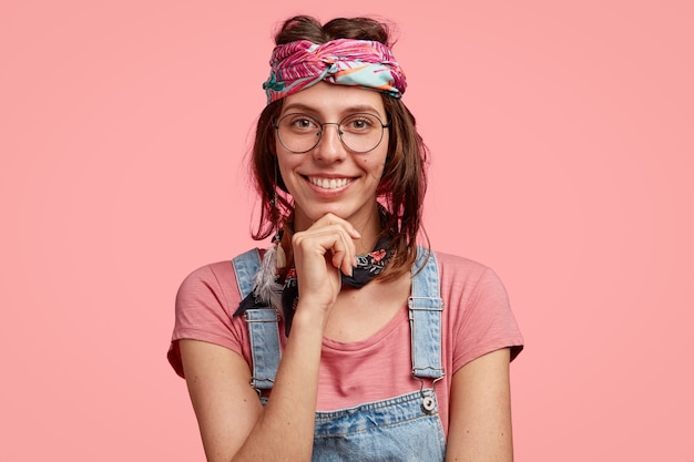 Foto gratuita foto de estudio de atractiva mujer hippie sostiene la barbilla, tiene una amplia sonrisa, lleva gafas redondas