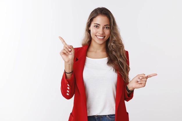 Foto de estudio atractivo amable sonriente feliz 25s mujer caucásica vistiendo chaqueta roja apuntando hacia los lados en diferentes direcciones sonriendo presentando opciones sonriendo sugiriendo bienes, pared blanca