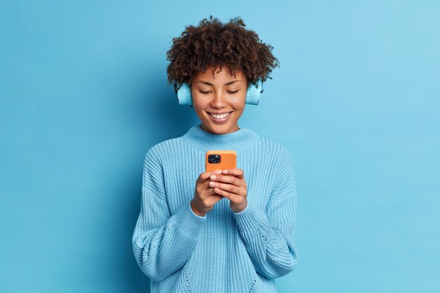 Foto de estudio de buena mujer milenaria de pelo rizado con amplia sonrisa utiliza el teléfono móvil para la comunicación en línea lleva auriculares estéreo elige una canción para escuchar
