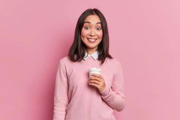 Foto de estudio de una chica asiática bastante morena tiene una pausa para el café sonríe suavemente a la cámara muestra dientes blancos vestidos con poses de puente casual en estudio