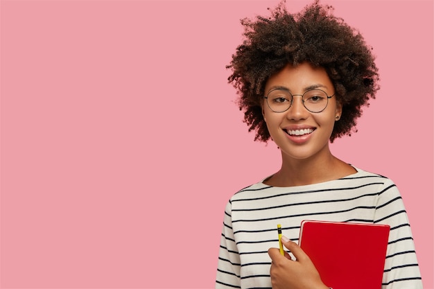 Foto de estudio de chica de piel muy oscura con una sonrisa suave, se prepara para las clases, lleva el bloc de notas rojo y lápiz