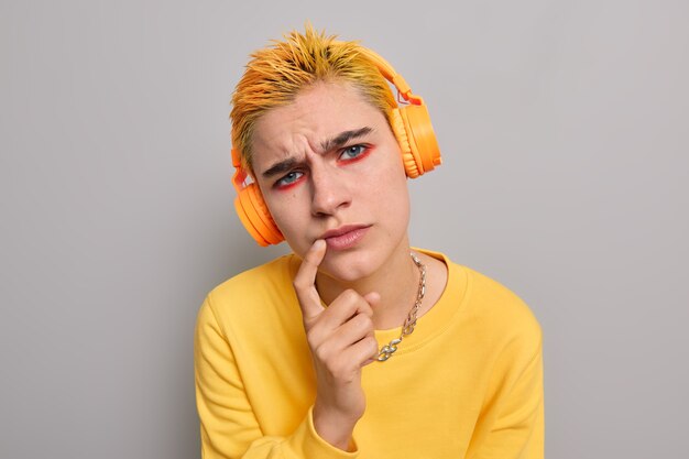 Foto de estudio de una chica punk atenta con peinado amarillo, maquillaje brillante, que mantiene el dedo índice cerca de los labios, intenta ver algo, escucha información con expresión concentrada, vestida de manera informal, posa interior