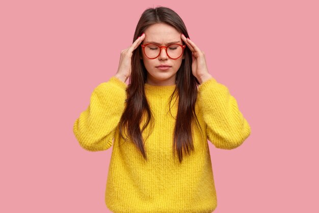 Foto de estudio de concentrada joven morena frota las sienes, tiene dolor de cabeza, se siente con exceso de trabajo