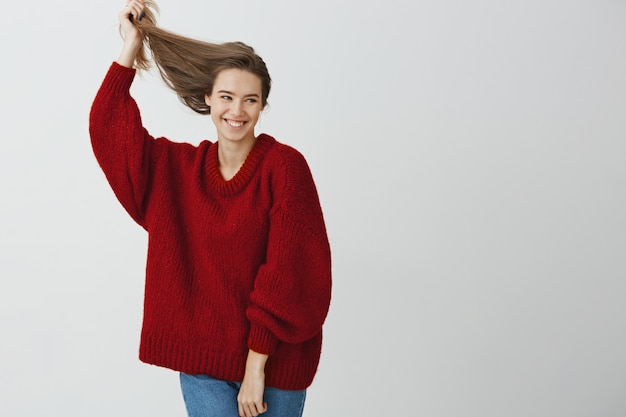 Foto gratuita foto de estudio de despreocupada mujer europea atractiva con una sonrisa sensual, levantando un cabello hermoso y saludable con la mano, mirando a un lado y expresando una actitud tierna y relajada, de pie sobre la pared gris