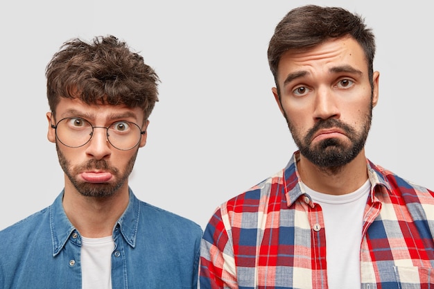 Foto gratuita foto de estudio de dos hombres guapos con barba que sienten insulto, labios curvados, tienen expresiones disgustadas, se sienten ofendidos por algo, posan contra la pared blanca